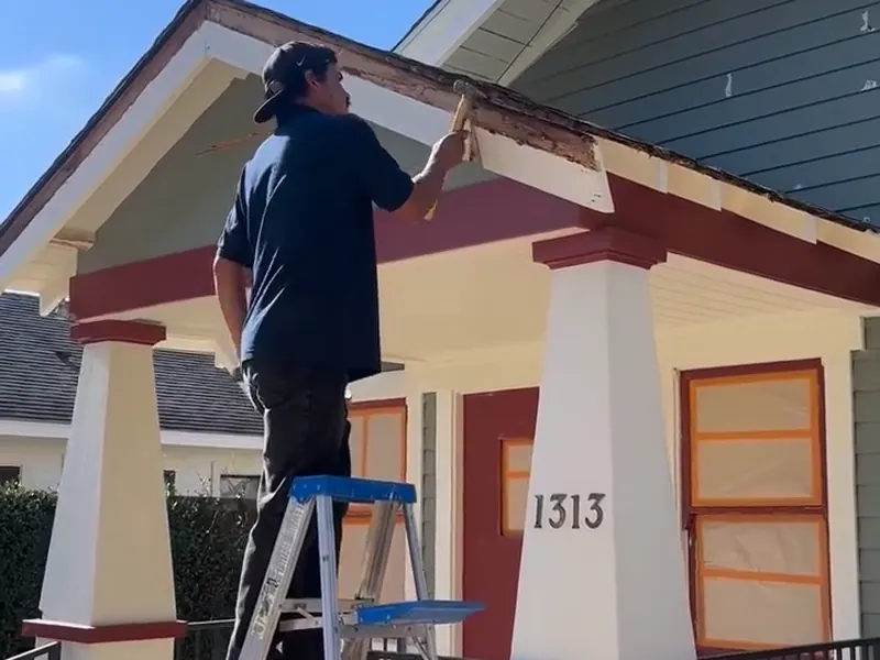 Santos worker scraping old paint from porch trim, with the windows masked for painting